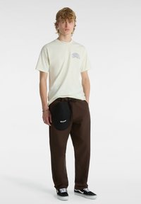 Lys beige t-shirt med et blåt grafisk mønster på venstre bryst, parret med brune bukser og en sort kasket, iført sorte og hvide sneakers.