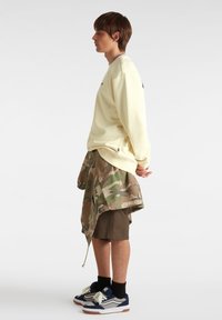 Lichtgele sweatshirt, camouflage wikkelrok over bruine shorts en marineblauwe sneakers met witte accenten. Casual gelaagde streetwear-stijl.