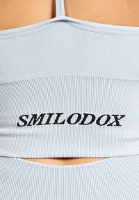 Sujetador deportivo de color azul claro con canalé, tirantes finos y un detalle recortado, que presenta el texto "SMILODOX" en letras negras en negrita.