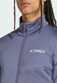 Terrex jakna na zip u svijetlo ljubičastoj boji, s visokim ovratnikom, mekanom teksturom i bijelim izvezenim logotipom na prsima.