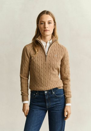 Frau trägt einen braunen, kabelgestrickten Halb-Reißverschluss-Pullover über einem weißen Hemd und blauen Jeans, stehend vor einem einfarbigen beigen Hintergrund.
