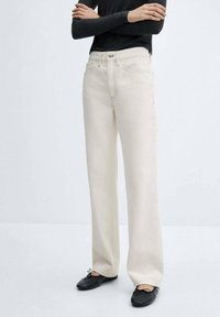 Jeans de pierna ancha color crema, hechos de mezclilla con costuras visibles y bolsillos frontales; combinados con zapatos negros.