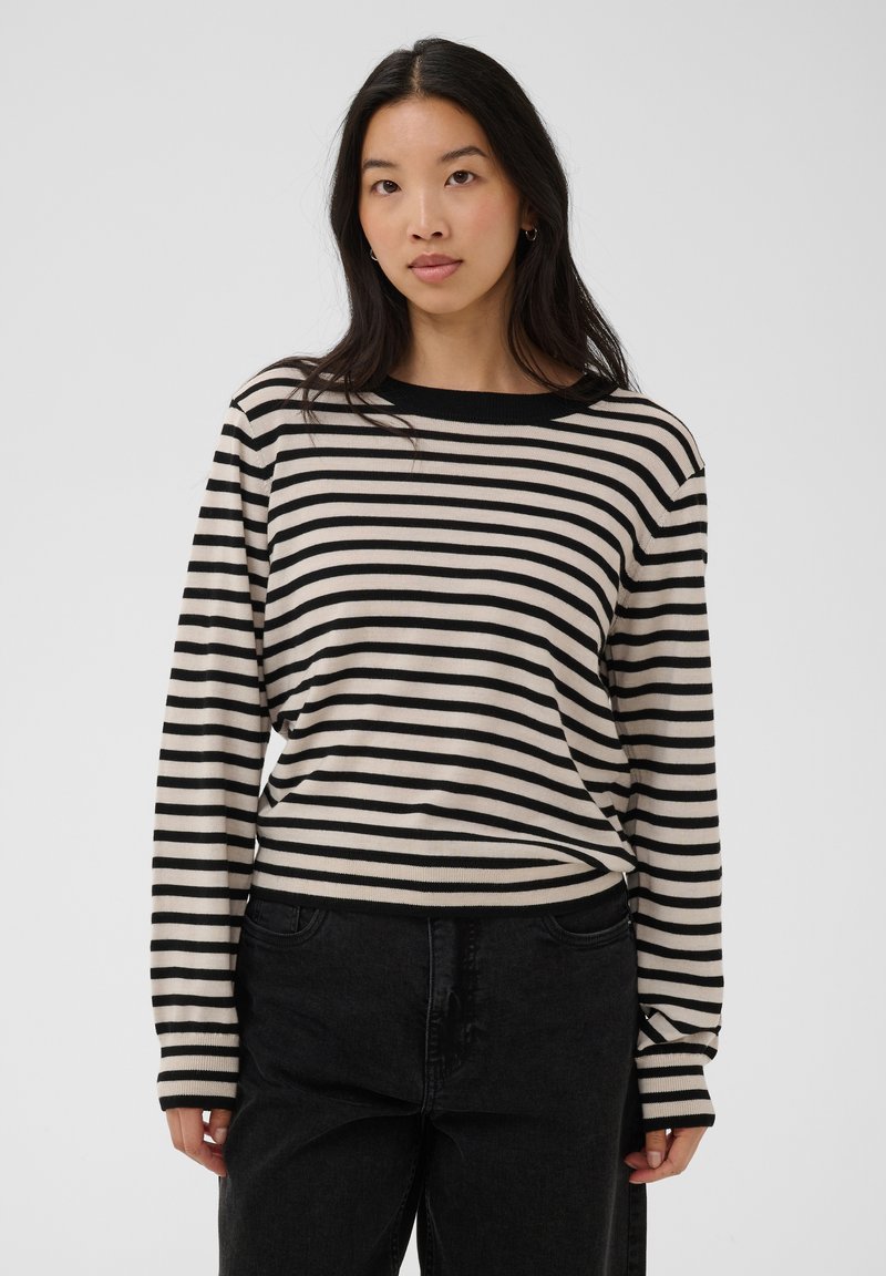 Femme aux longs cheveux noirs portant un haut à manches longues rayé noir et blanc et un pantalon noir, debout devant un fond clair uni.
