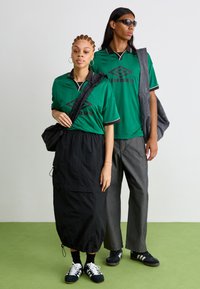 Polo verde de manga curta com detalhes em preto, combinado com uma saia curta preta. Modelo a usar ténis pretos e uma mochila de cintura. Calças cinzentas.