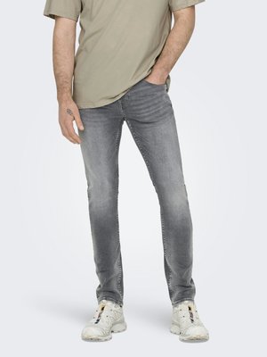 Άνδρας φορώντας ξεθωριασμένο γκρι slim-fit τζιν, μπεζ μπλουζάκι και λευκά αθλητικά παπούτσια με πολύχρωμες λεπτομέρειες, στέκεται με το ένα χέρι στην τσέπη.