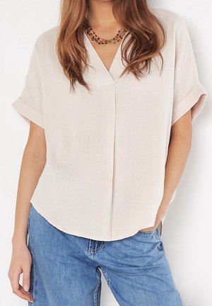 Femme portant un chemisier beige ample à manches courtes avec col en V et manches retroussées, associée à un jean bleu et un collier multicolore en perles.