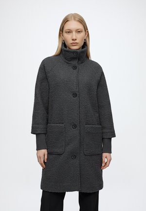 Femme portant un manteau texturé gris foncé longueur genoux avec de grandes poches avant et un col haut boutonné, debout devant un fond uni.