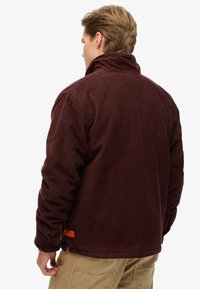 Bordeaux ribfluwelen jas met een hoge kraag, ritssluiting en ribbelstructuur. Heeft een oranje logo label dicht bij de zoom.
