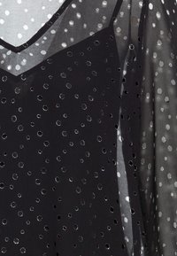 Schwarzes transparentes Oberteil mit V-Ausschnitt, kreisförmigen Ausschnitten und metallischen Akzenten, die ein strukturiertes Muster im gesamten Stoff erzeugen.