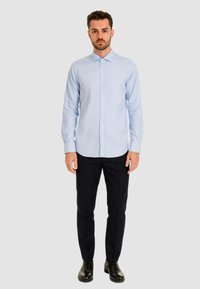 Camicia azzurra a maniche lunghe con colletto appuntito, abbinata a pantaloni scuri e scarpe nere, su uno sfondo neutro.