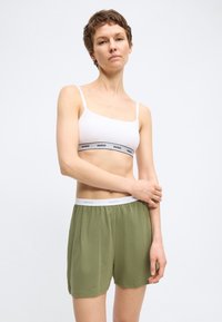 Γυναίκα που φοράει λευκό crop top με λογότυπο Hugo και πράσινα σορτς με ελαστική μέση και λογότυπο Hugo, στέκεται μπροστά σε απλό φωτεινό φόντο.