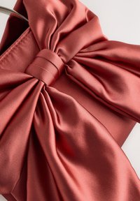 Rožnata satenasta tkanina z velikim, strukturiranim lokom na vrhu, z gladko teksturo in natančno zbranimi gubami, pritrjenimi s centralnim trakom.