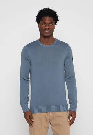 Uomo con capelli corti e ricci che indossa un maglione di lana blu e pantaloni beige, in piedi davanti a uno sfondo chiaro e uniforme.