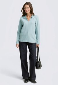 Lichtblauw shirt met lange mouwen en een kraag, met een v-hals ontwerp. Draagt donkere bootcut jeans en een kleine zwarte crossbodytas.