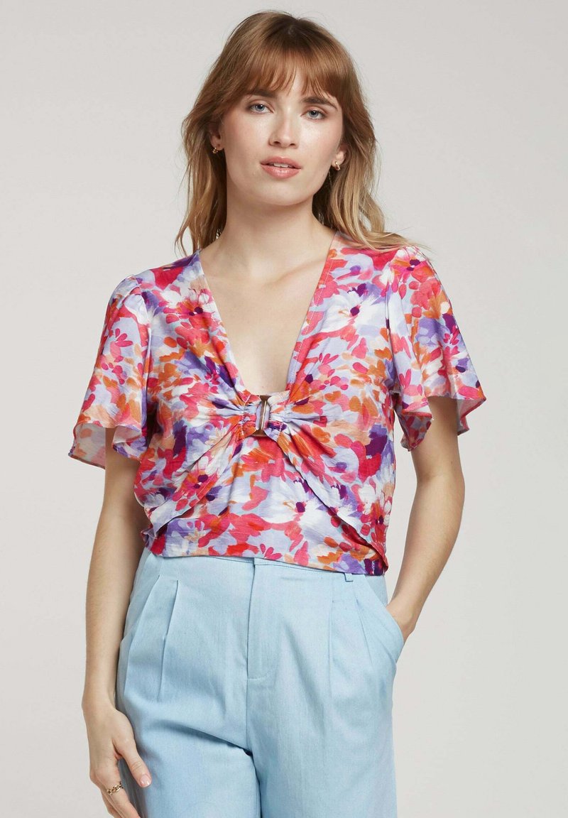 Blouse fleurie avec des manches courtes en volant, dotée d'un détail à nouer à la taille et de couleurs vives de rose, orange et violet ; tissu léger.