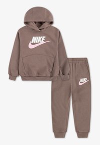 Hnedá mikina s kapucňou a zodpovedajúce tepláky, s ružovým logom Nike. Vyrobené z jemného materiálu s prúžkovanými manžetami a pásom.