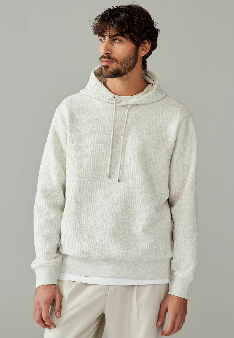 Lys grå tekstureret hoodie med en snoretrækshætte, ribbede manchetter og kant, parret med et hvidt underlag. Casual pasform.