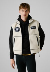 Prsluk od flisa u boji off-white s crnim detaljima i akcentima. Ima bočne džepove i izvezene zakrpe. Nosi se preko crnog dukserice i traperica.
