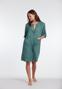 Groene jurk tot de knie, gemaakt van een gladde stof, met een ingesneden kraag, opgerolde mouwen en twee voorzakken voor opslag.