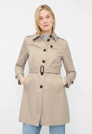 Frau trägt einen beigen Trenchcoat mit Gürtel und schwarzen Knöpfen über blauer Jeans, steht mit den Händen in den Taschen vor weißem Hintergrund.