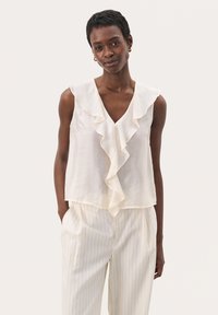 Ivory sleeveless blouse with a V-neckline and front ruffles; made of lightweight fabric. Paired with cream-striped trousers.