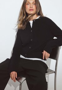 Melnā kardigāna ar ribotu tekstūru, V-veida kaklu, sešām pogām un garām piedurknēm, pārī ar baltu kreklu un tumšiem biksēm.