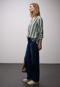 Femme debout de profil portant un chemisier rayé à manches longues, un jean large bleu, des baskets blanches, tenant un sac marron.