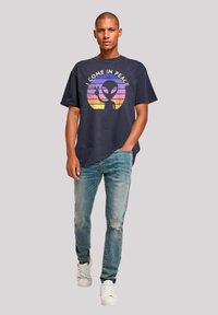 Navy cotton t-shirt with round neckline featuring a graphic of an alien and the text "I COME IN PEACE." Worn with light blue distressed jeans and white sneakers.