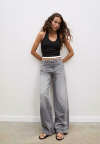 Jeans grigi ampi con sfumature, a vita alta, abbinati a un top corto nero. La modella è in piedi di fronte a uno sfondo semplice.