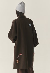 Barna gyapjúkabát túlméretezett ujjal és hímzett részletekkel: egy madár a hátán és egy gyerek sárkányt reptet az oldalán. Texturált anyag.