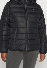 Černá prošívaná bunda s kapucí, s vodorovně prošívanými sekcemi, s zipem a bočními kapsami. Vyrobena z lesklého, lehkého materiálu.