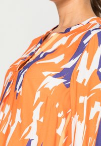 Oranžová a bílá košile s abstraktními štětcovými vzory a fialovými akcenty, s kulatým výstřihem a uvolněným střihem.