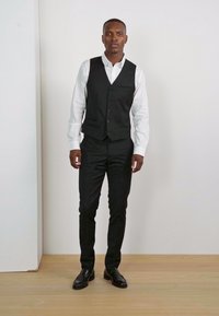 Schwarze Weste über einem weißen Hemd, schwarze Hose und schwarze Schuhe. Die Weste hat einen Knopfverschluss und eine Brusttasche. Minimalistisches Design.