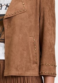 Veste en daim marron avec des accents cloutés, dotée d'un col cranté, de poches latérales et de manches détaillées ornées de clous dorés. La texture est douce et lisse.