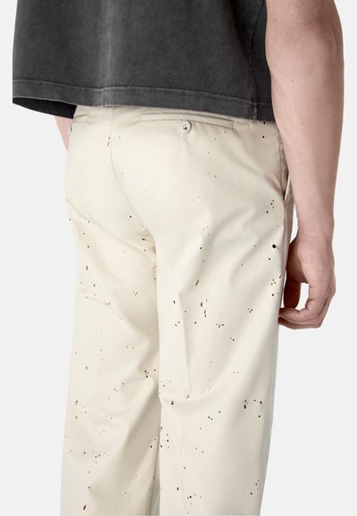 Pantalones beige con patrón salpicado oscuro, usados por una persona con una camiseta corta gris oscuro, mostrados desde la cintura hacia abajo, vista trasera.