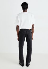 Jeans de mezclilla negra con un corte recto. Presentan cinco bolsillos, costuras visibles y una etiqueta de tela en la parte trasera de la cintura. Combinados con una camiseta blanca.