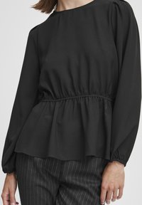 Blusa nera a maniche lunghe con scollatura rotonda, vita arricciata e orlo peplum fluido. Tessuto morbido con una leggera lucentezza. Abbinata a pantaloni a righe. 