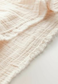 Světle růžová bavlněná mušelínová látka s texturovaným mřížkovým vzorem a třásněmi, rozprostřená na bílé ploše.