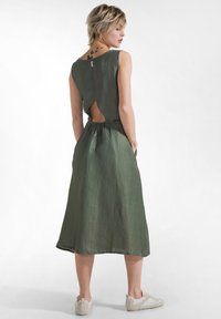 Olivgrön ärmlös klänning med utskuren rygg, markerad midja och flödande fåll. Tillverkad av lätt, strukturerat tyg. Vita sneakers.