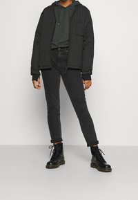 Svart vadderad bomberjacka med ribbade muddärmar, lager-på-lager över en mörkgrå hoodie. Matchad med svarta slim-fit jeans och svarta snörkängor.