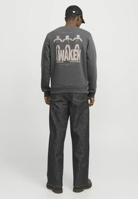 Mørkegrå sweatshirt med grafisk tryk "AWAKEN" og tre figurer på ryggen. Bæres med løstsiddende mørke jeans og sort kasket.
