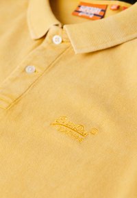 Polo giallo con una trama texturizzata, dotato di colletto, tre bottoni e un logo ricamato in una tonalità di giallo abbinata.