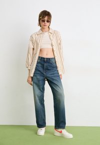 Camisa de botón a rayas beige sobre un top crema corto, combinada con jeans azules sueltos y zapatillas blancas con acentos rojos. Fondo de suelo verde.