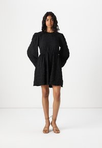 Vestido negro corto con tejido texturizado, mangas largas abullonadas y un corsé ajustado con falda acampanada. Sandalias planas completan el look.