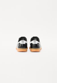Sneakers de couro preto com um logotipo branco, apresentando uma sola de borracha de goma, painéis laterais texturizados e um design de perfil baixo.