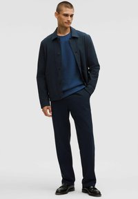 Joven vestido con una chaqueta azul marino, un suéter azul, pantalones oscuros y zapatos negros, de pie con una mano en el bolsillo.