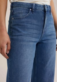 Närbild av en person som bär blå denimjeans med en knapp och dragkedja, som visar framfickor och en del av en hand som vilar vid sidan.