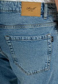 Jeans de mezclilla de color azul claro con una etiqueta de cuero que dice "Reell est. 1997" en la parte trasera, con un único bolsillo trasero y costuras en contraste.
