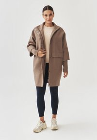 Bruine wolmix jas met een capuchon, voordeknopen en zijzakken, gedragen over een lichte trui en donkere leggings, gecombineerd met witte sneakers.