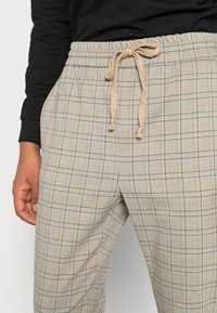 Light beige checkered trousers with a drawstring waist, featuring a subtle plaid pattern in green and blue. Soft fabric texture.
